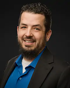 Head-and-shoulders portrait of a smiling man with dark hair and beard, wearing a blue shirt and black blazer against a dark backdrop.